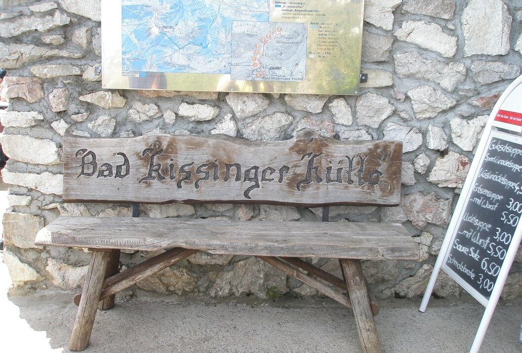 Holzbank "Bad Kissinger Hütte" vor Steinmauer mit Wanderkarte