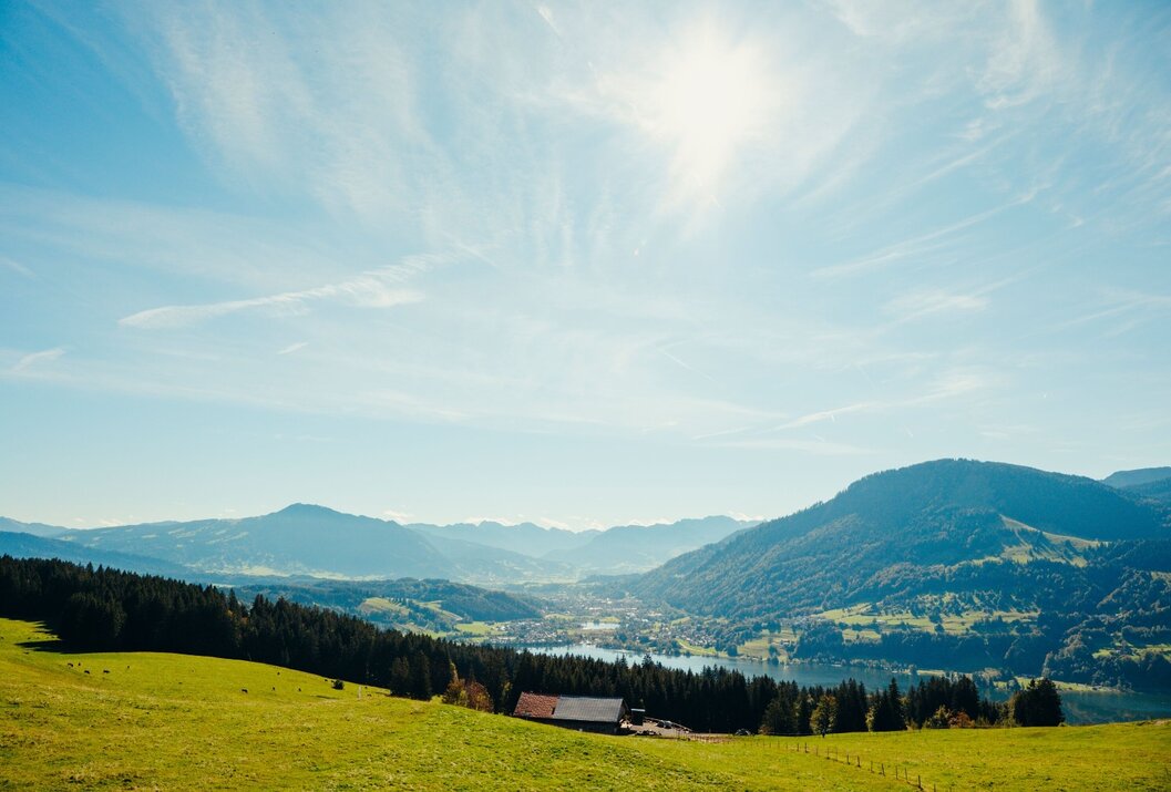 Hügelige Landschaft mit Wiesen, Wald, Bergen, See und einem kleinen Haus unter blauem Himmel mit Sonne