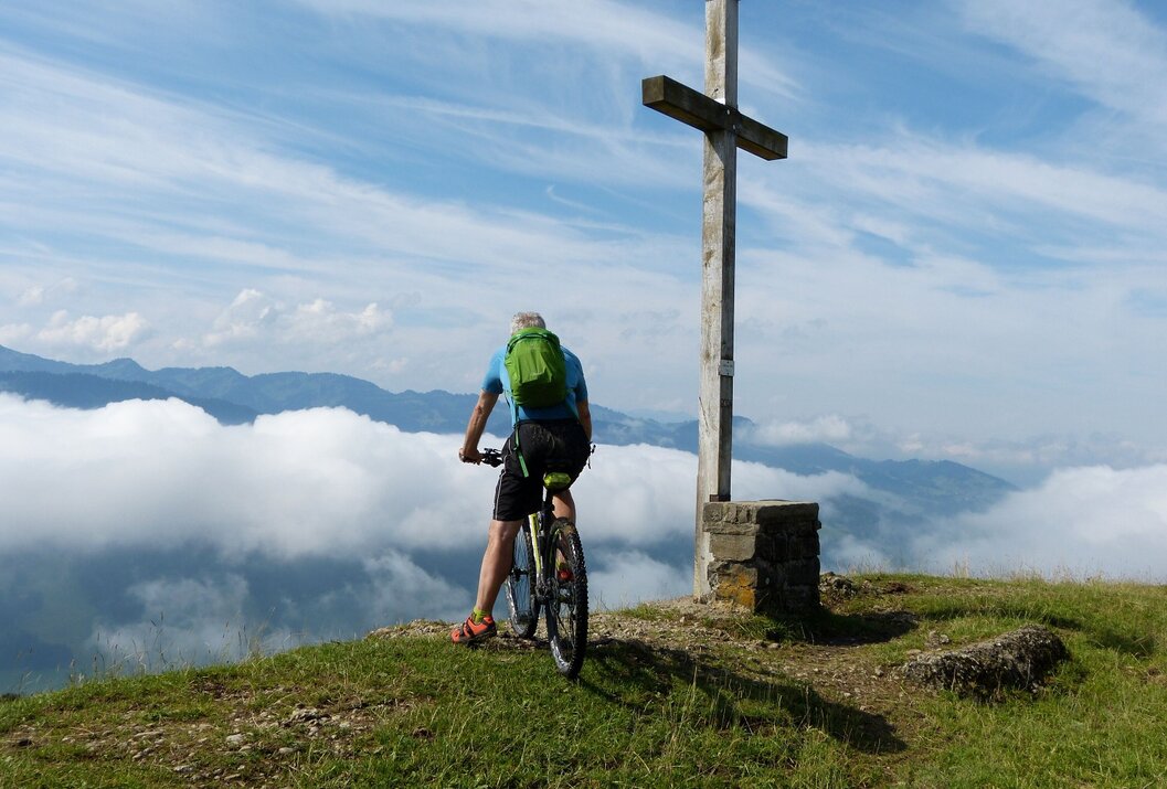 Mann mit Mountainbike vor Gipfelkreuz mit Wolkenmeer im Hintergrund
