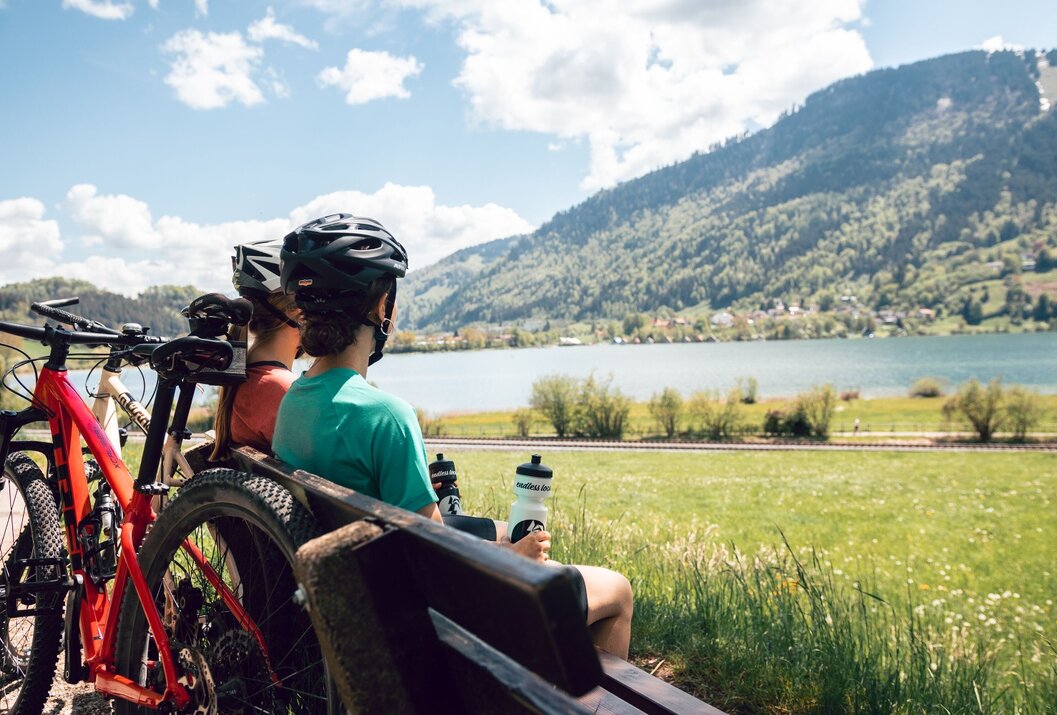 Zwei Radfahrer rasten auf Bank mit Blick auf See und Berge