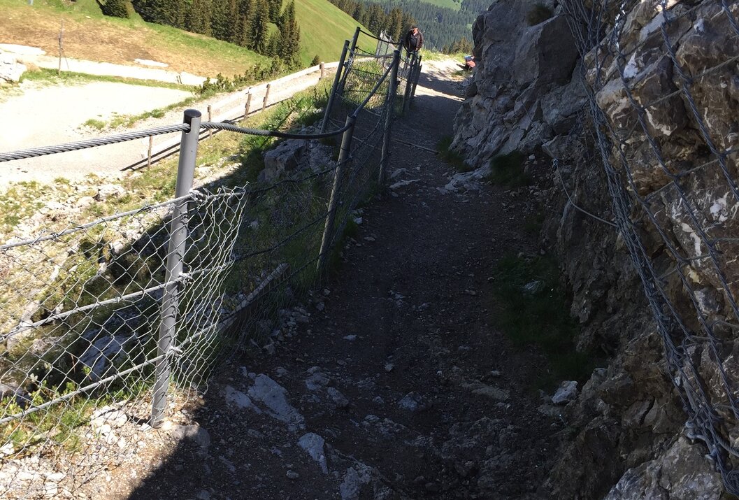 Schmaler Bergpfad mit Zaun neben Felswand, im Hintergrund grüne Hügel