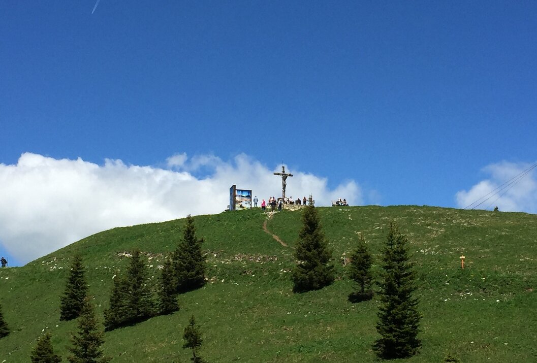 Gipfelkreuz auf grünem Hügel unter blauem Himmel mit Wolken