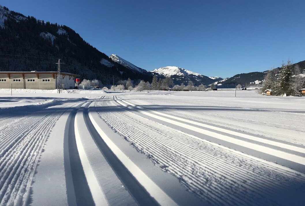 Verschneite Langlaufloipe vor Bergkulisse unter blauem Himmel