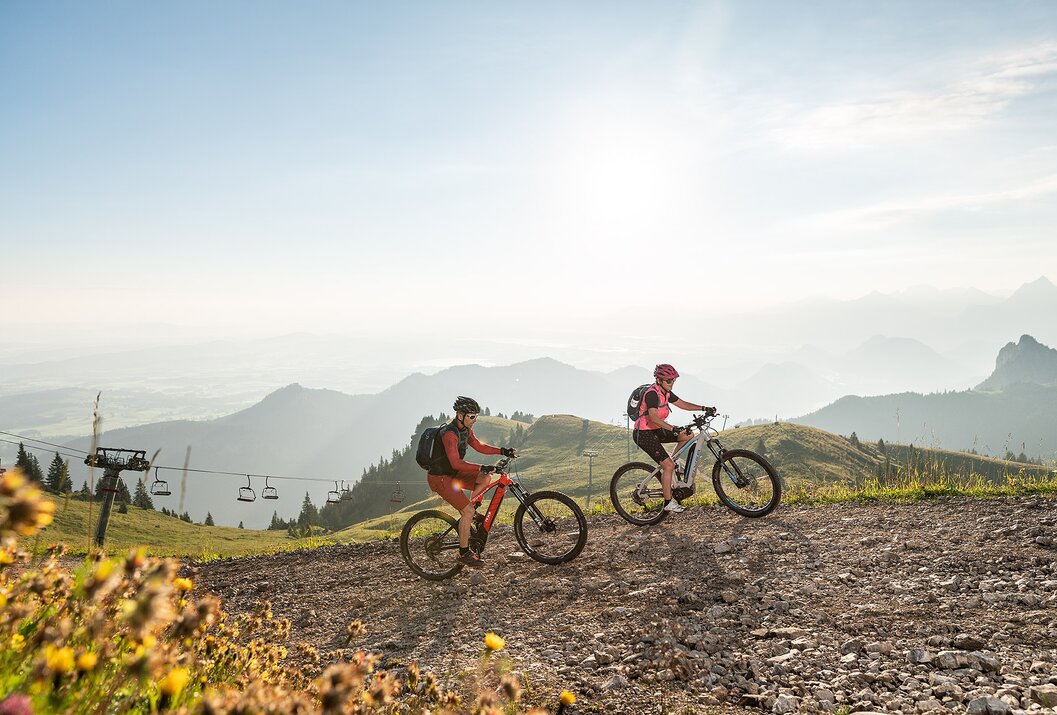 Zwei Mountainbiker fahren bergauf auf einem Schotterweg vor Bergpanorama