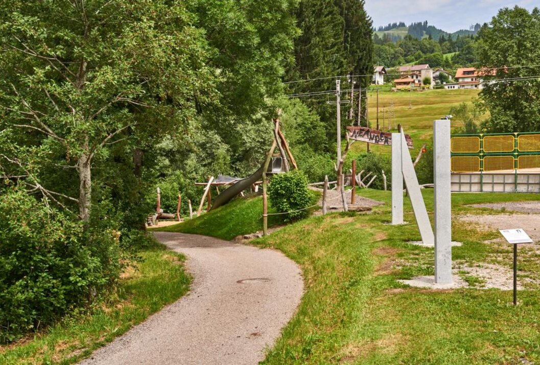 Geschwungener Weg führt zu Spielplatz mit Holzhäuschen und weißen Pfosten vor grüner Landschaft