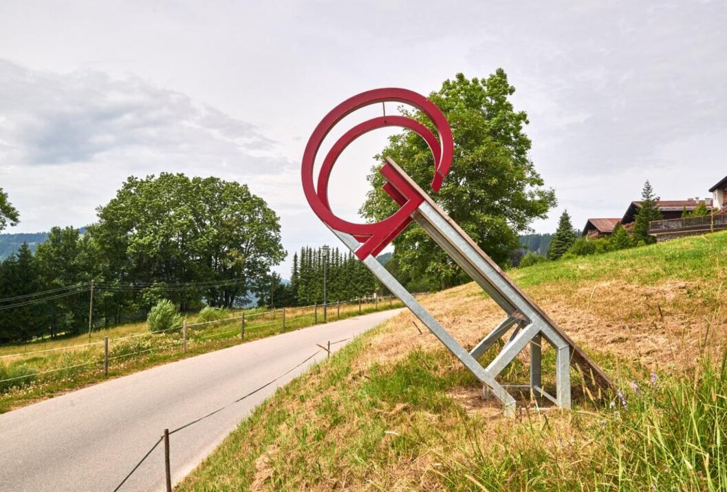 Rote Skulptur mit zwei Kreisen auf Metallgestell an einem Hang neben einer Straße