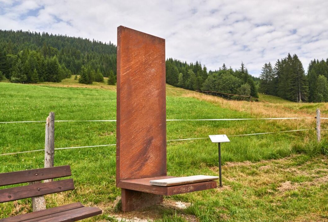 Rostfarbene Metallskulptur mit Sitzfläche auf grüner Wiese vor bewaldetem Hügel