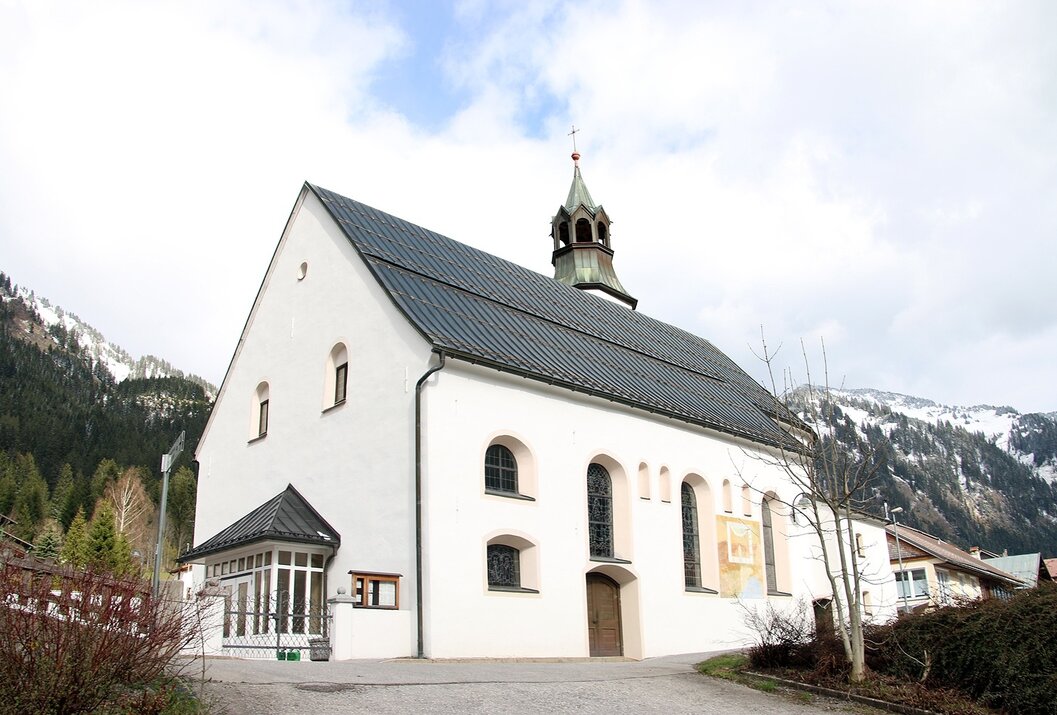 Weißes Gebäude mit dunklem Dach und kleinem Turm vor bewaldetem Hügel
