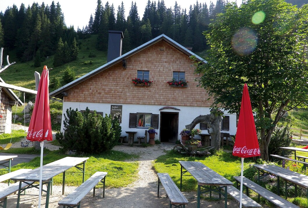 Hütte mit Holzdach und weißen Wänden vor Wald, Tische und Bänke im Vordergrund