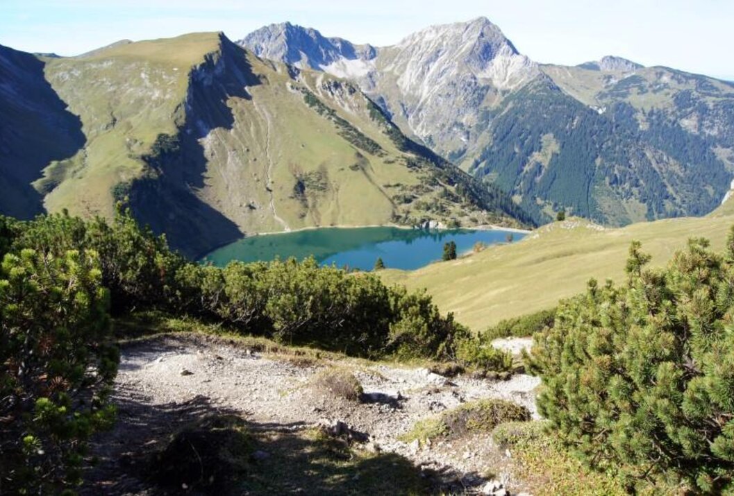 Bergsee eingebettet in grüne Hänge, im Hintergrund hohe, felsige Gipfel
