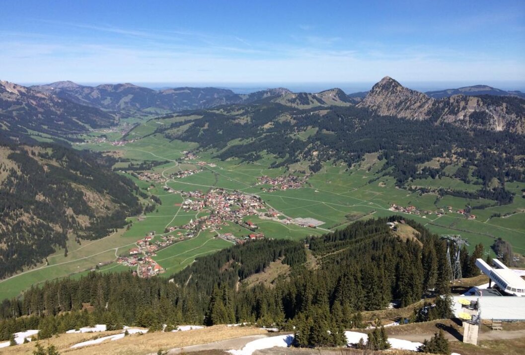 Panorama: Blick von Berg auf grünes Tal mit Dorf, umgeben von bewaldeten Hügeln unter blauem Himmel