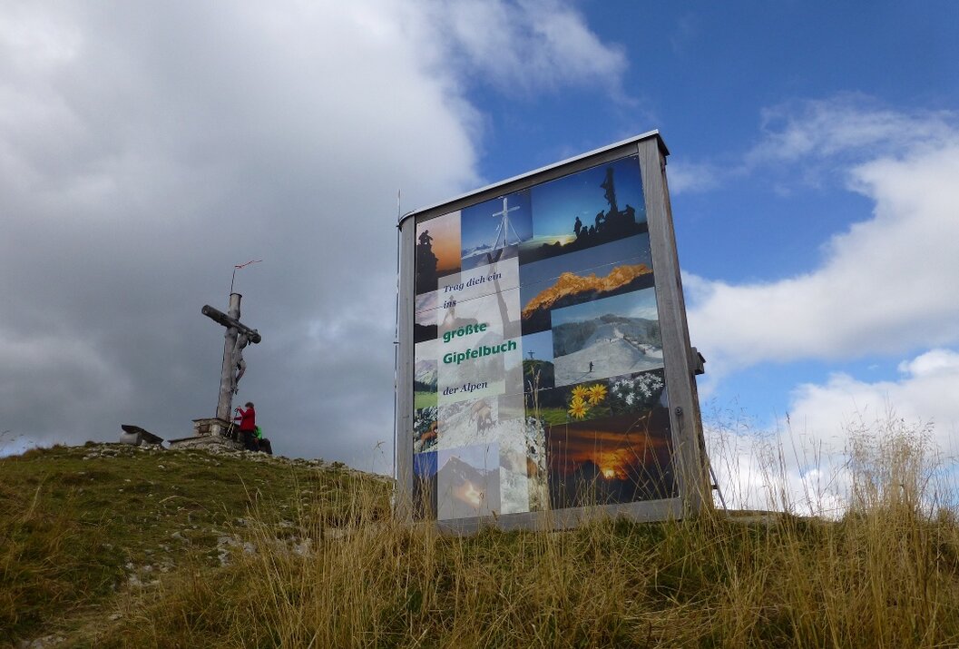 Gipfelkreuz und Gipfelbuch-Hütte auf Bergkuppe unter bewölktem Himmel