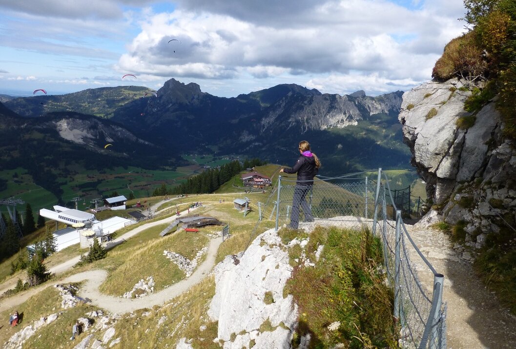 Frau steht an einem Felsvorsprung und blickt auf eine Berglandschaft mit Häusern und Gleitschirmfliegern