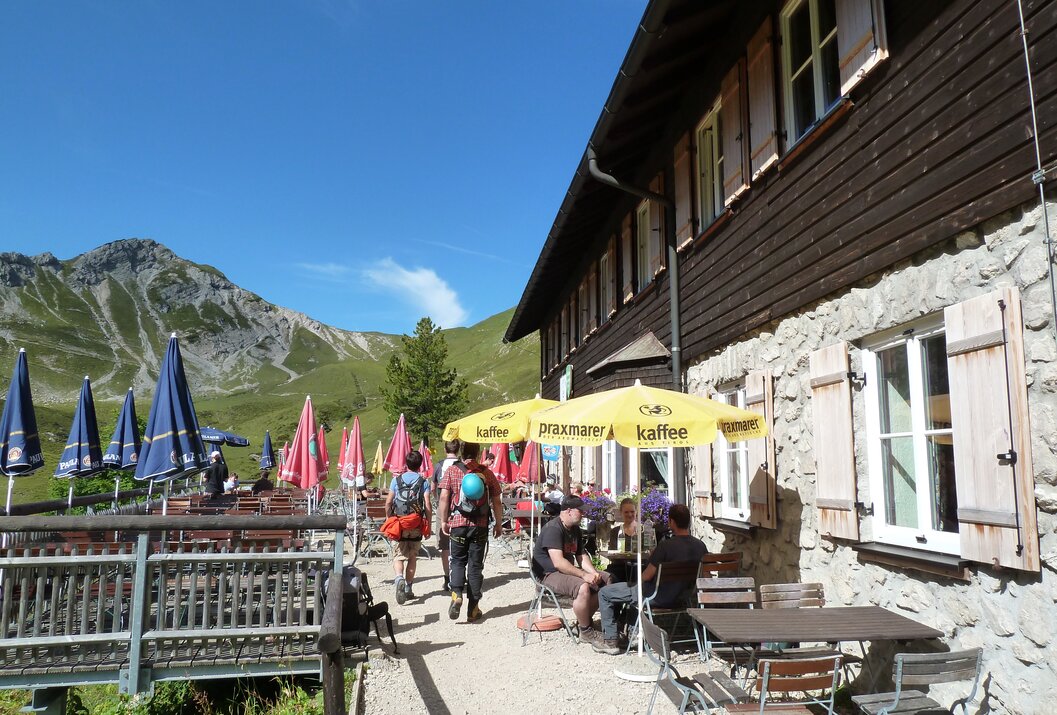 Berghütte mit Gästen auf Terrasse, Sonnenschirme, Berglandschaft im Hintergrund