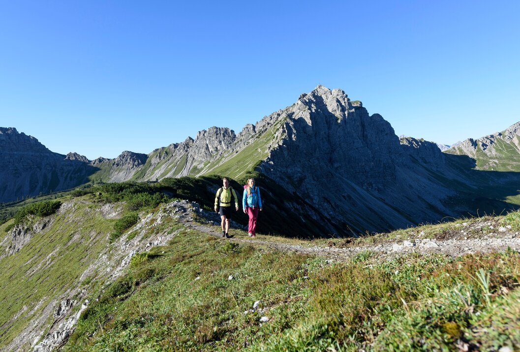 Zwei Personen wandern auf einem Bergpfad vor einer Bergkulisse