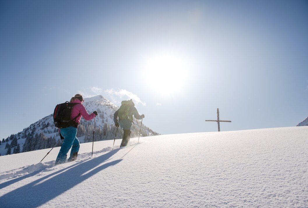 Zwei Personen steigen mit Skiern einen schneebedeckten Hang hinauf, im Hintergrund ein Gipfelkreuz und strahlender Sonnenschein