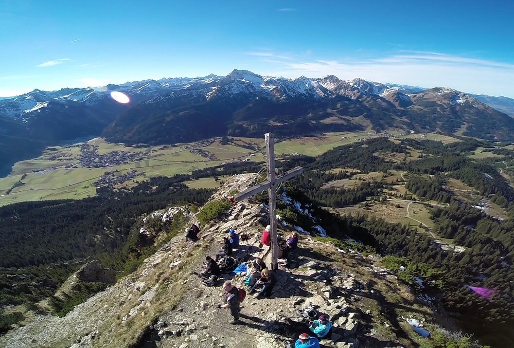Gipfelkreuz auf Bergspitze, Menschengruppe, bewaldete Landschaft und schneebedeckte Berge im Hintergrund
