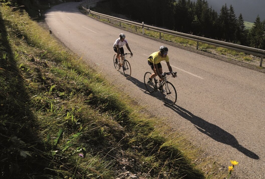 Zwei Radfahrer auf kurvenreicher Bergstraße vor bewaldeter Bergkulisse