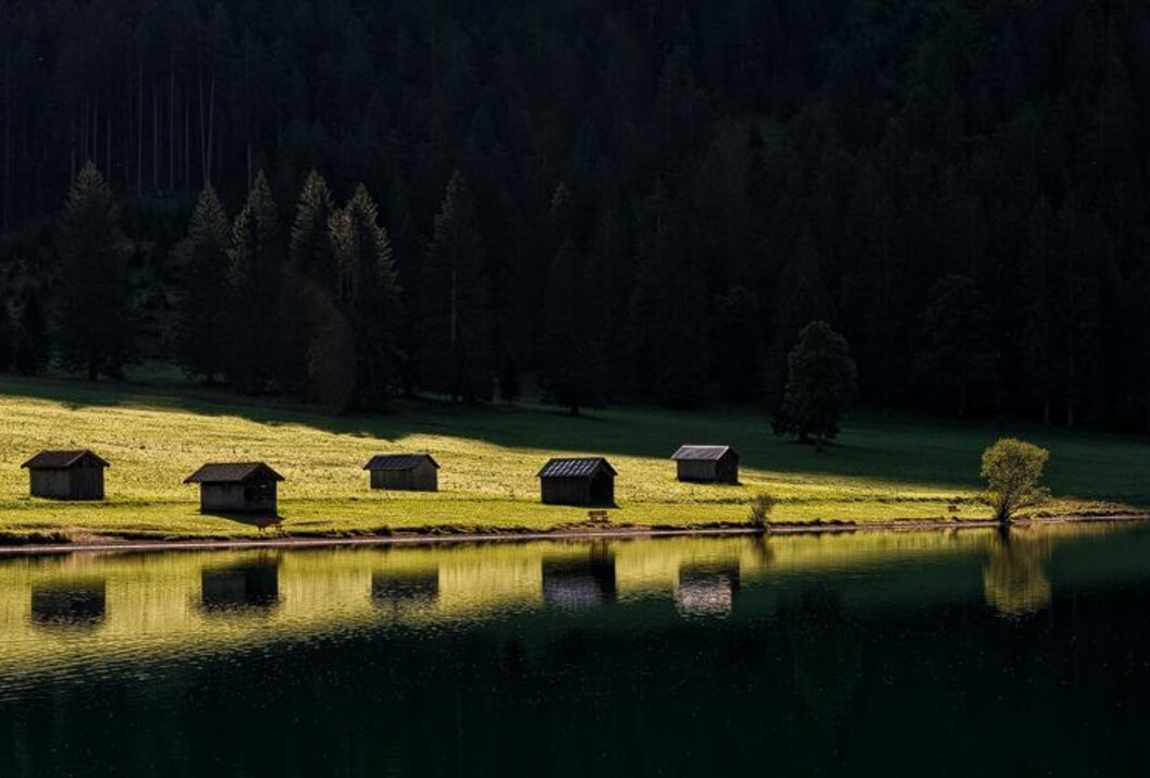 Vier kleine Hütten am Seeufer mit Spiegelung im Wasser vor dunklem Wald