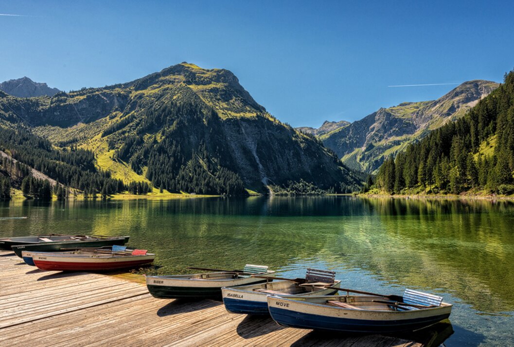 Bergsee mit Booten an einem Holzsteg vor bewaldeten Bergen unter blauem Himmel