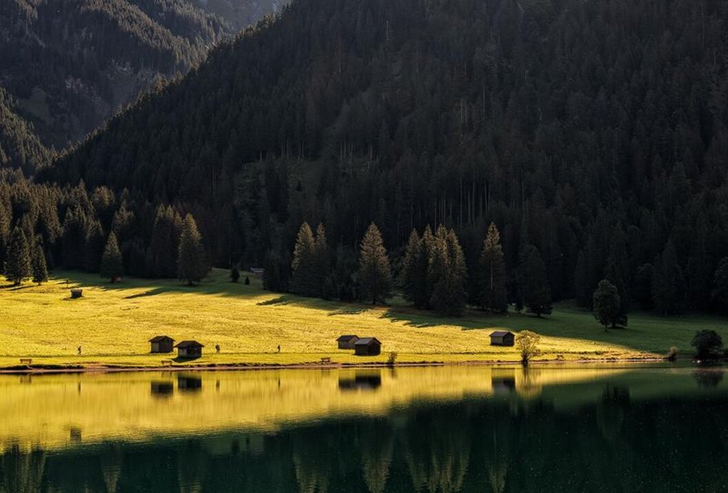 Idyllische Landschaft mit See, Wiese, Hütten und dunklem Wald im Hintergrund