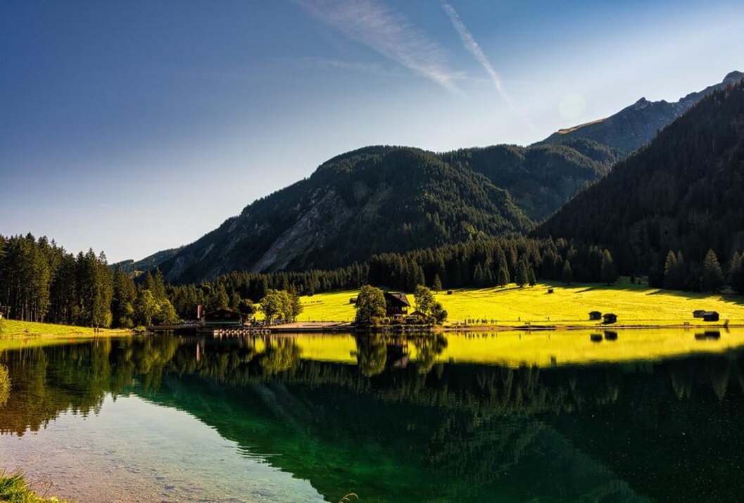 Bergsee mit grünen Wiesen, bewaldeten Bergen und Spiegelung im Wasser