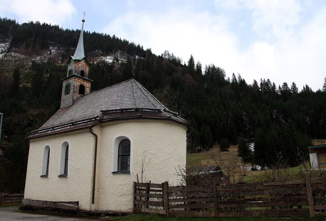 Kleine weiße Kirche mit grünem Turm vor dunklem Wald und bewölktem Himmel