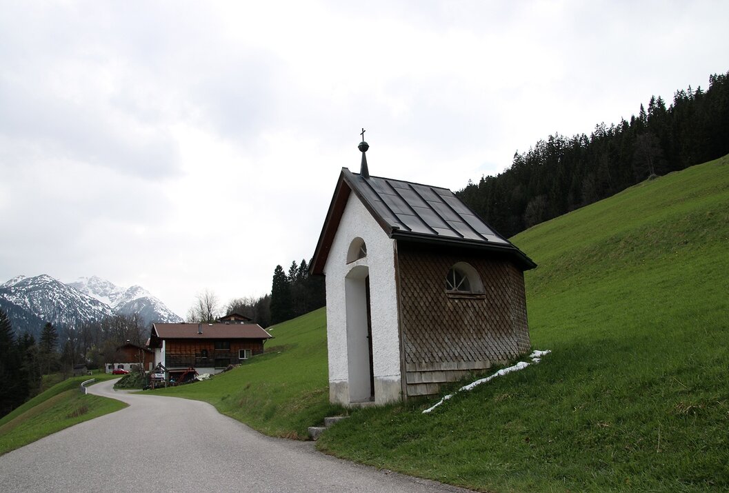 Kleine Kapelle am Wegrand vor grüner Wiese und Häusern