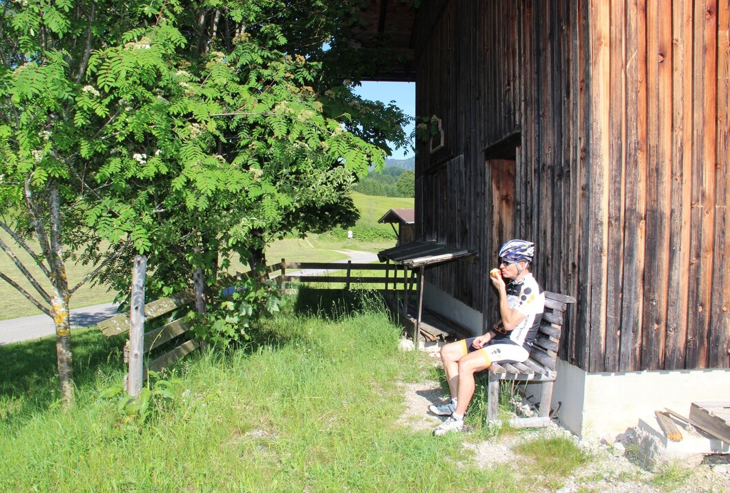 Radfahrer sitzt auf Bank vor Holzhütte, isst und blickt in grüne Landschaft