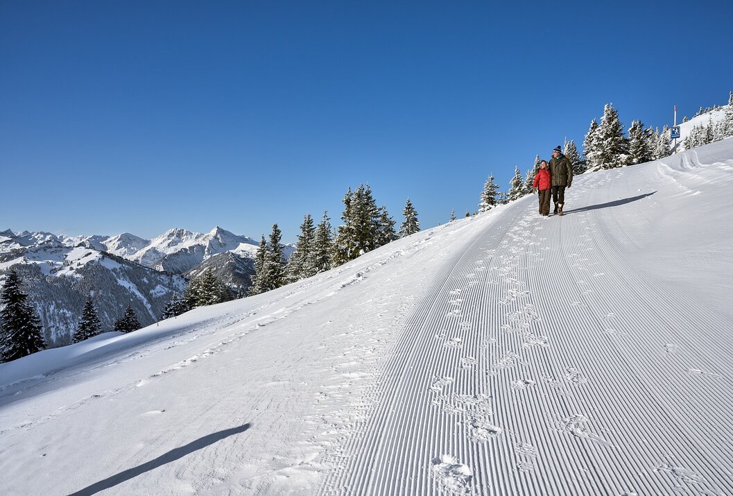 Paar wandert auf schneebedecktem Weg, im Hintergrund Berge und blauer Himmel