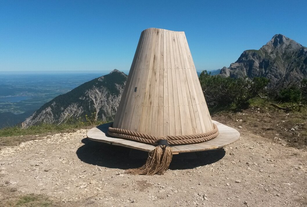 Hut-Skulptur aus Holz auf Bergspitze mit Seil und Quaste vor Bergpanorama