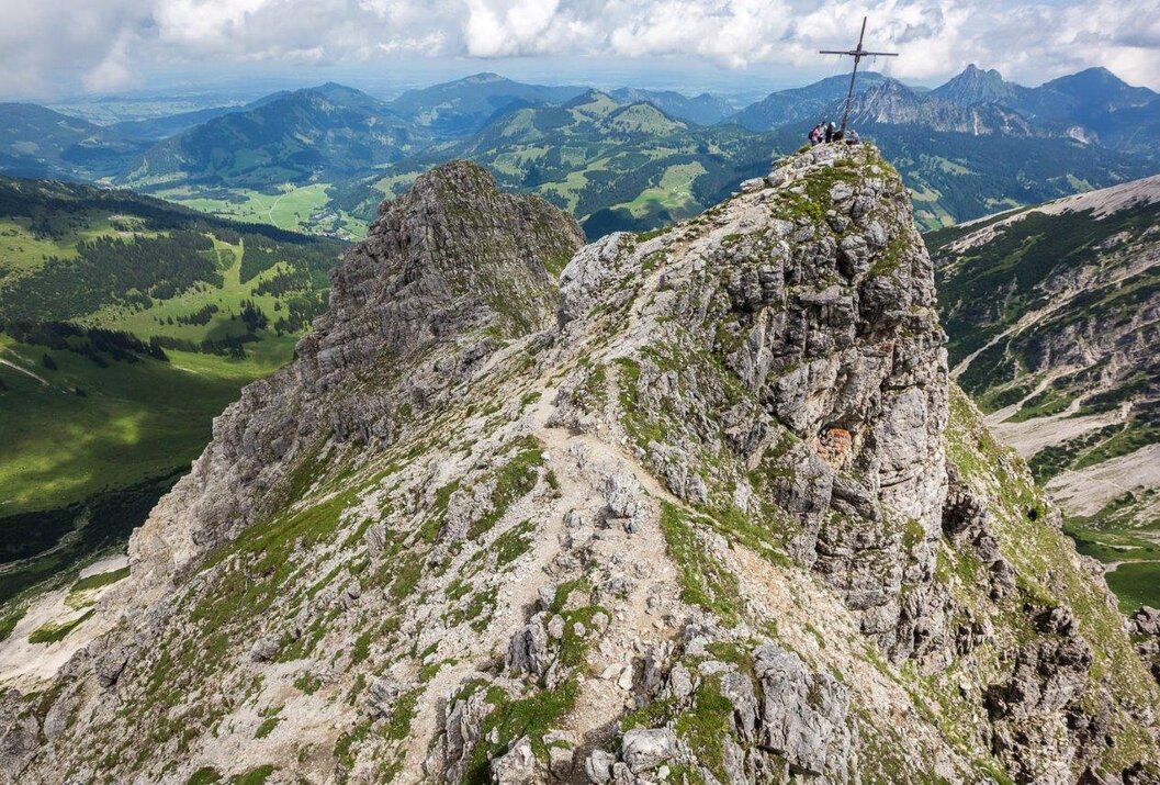 Bergpfad führt zu Gipfelkreuz mit Personen, grüne Hügel im Hintergrund