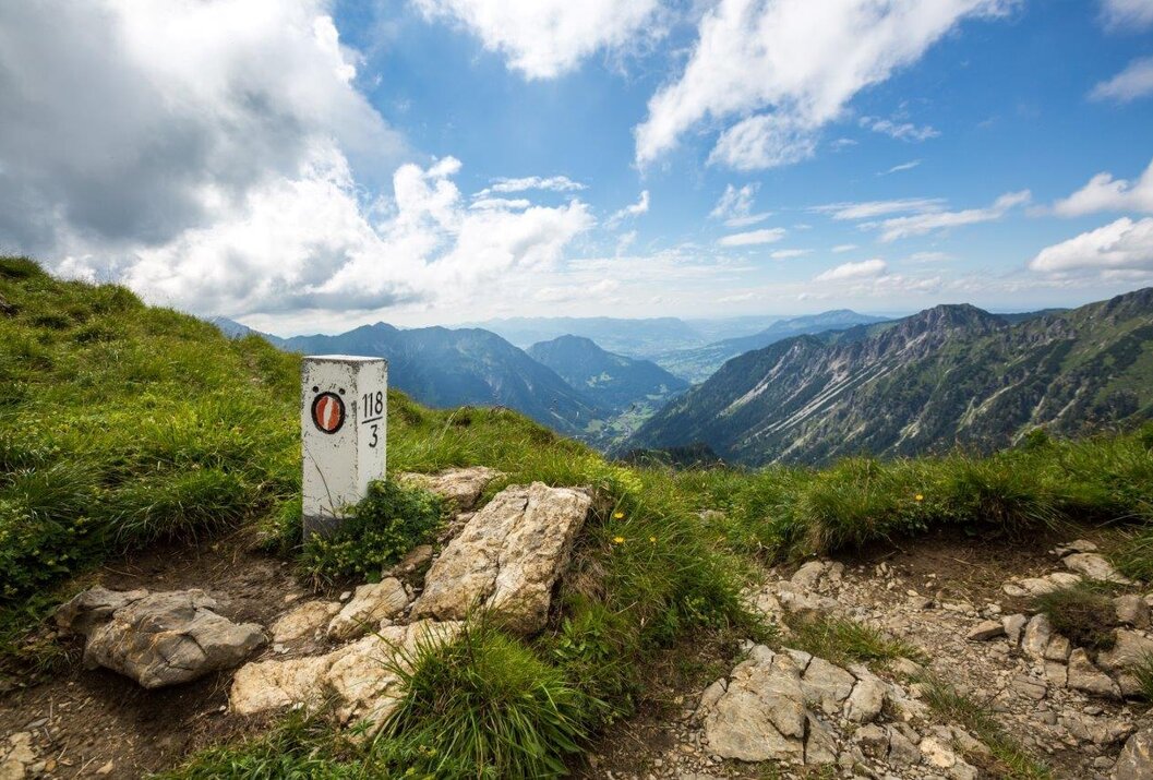 Wanderwegweiser auf Bergwiese mit Blick auf grüne Täler und bewölkten Himmel