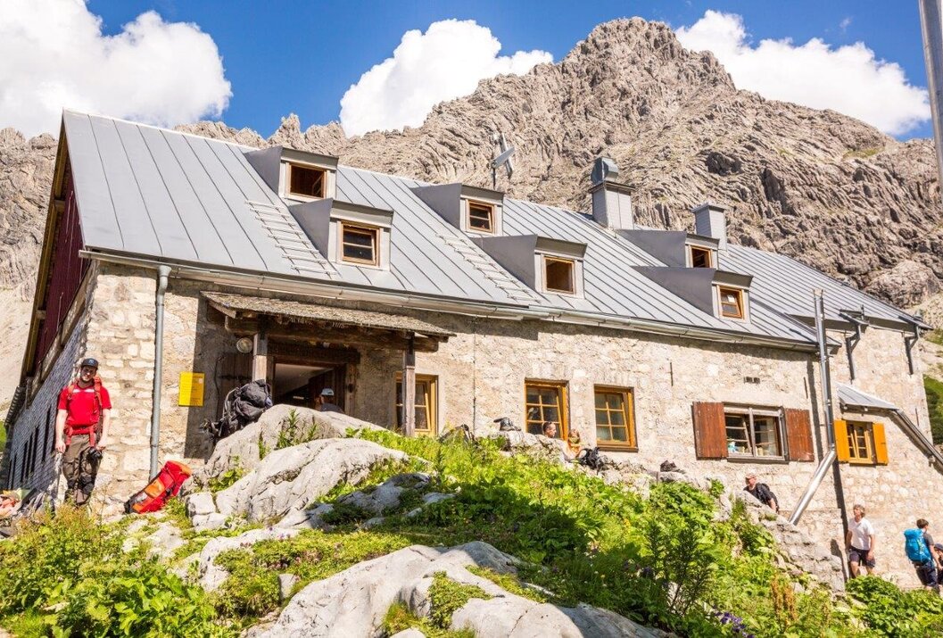 Berghütte aus Stein mit grauen Dach vor felsigem Berg unter blauem Himmel