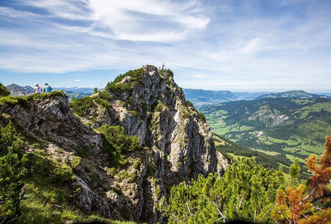 Bergspitze mit Gipfelkreuz vor grüner Landschaft unter blauem Himmel mit Wolken