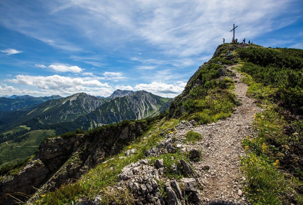 Bergpfad führt zu Gipfelkreuz mit Personen, im Hintergrund Bergpanorama unter blauem Himmel
