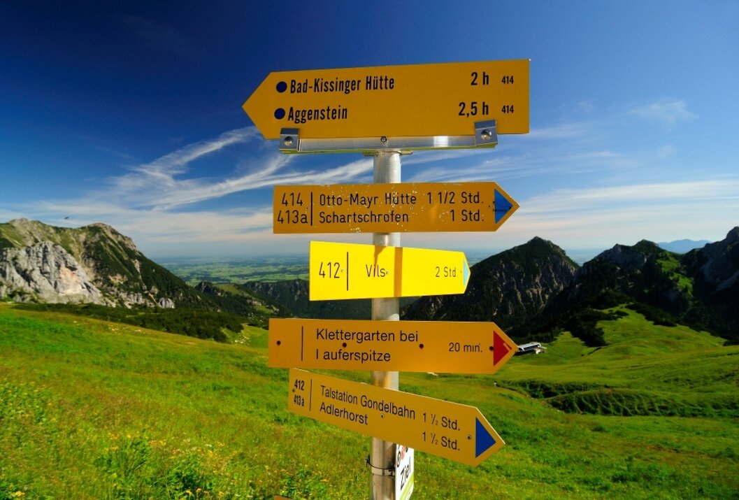 Gelbe Wegweiser mit Wanderzielen vor Berglandschaft und blauem Himmel