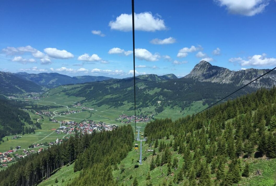 Blick aus einer Seilbahn auf grüne Hügel, Berge und ein Dorf unter blauem Himmel mit Wolken