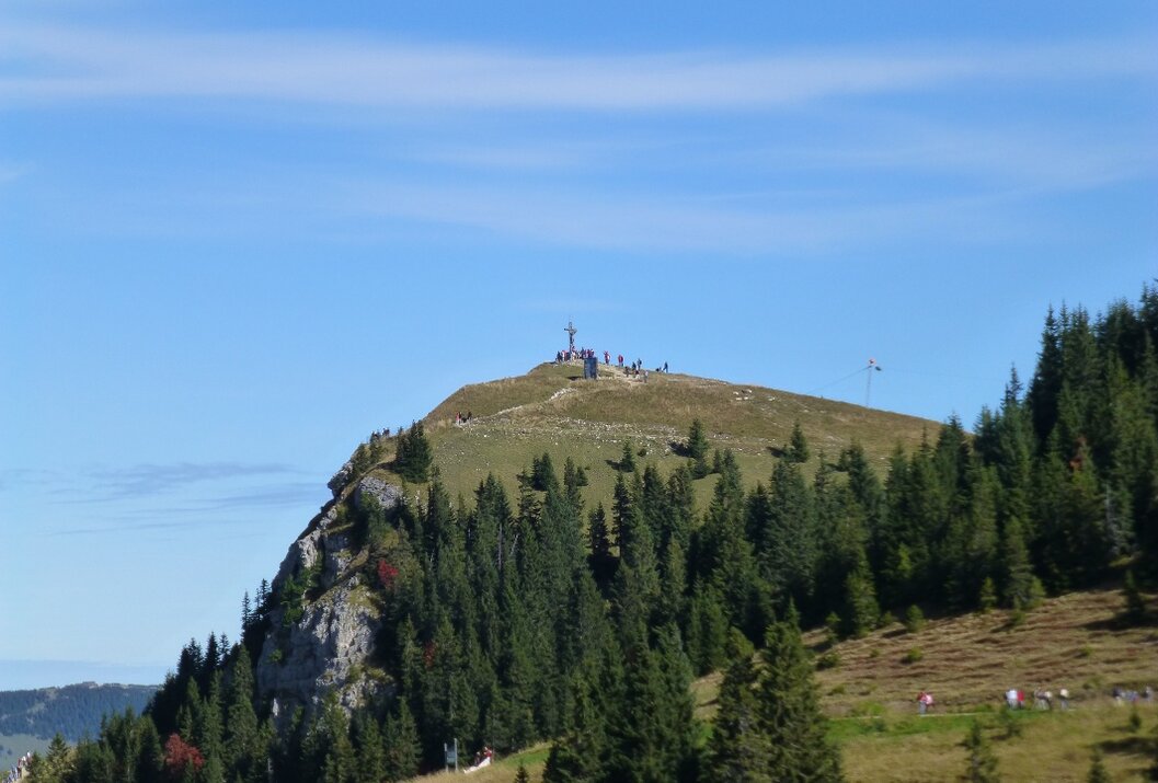 Bergspitze mit Kreuz und Menschen unter blauem Himmel, umgeben von Bäumen