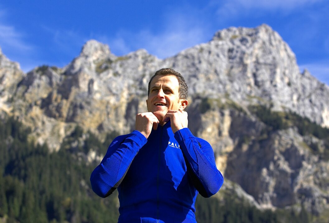 Mann in blauem Oberteil vor Bergkulisse fasst sich an den Kragen