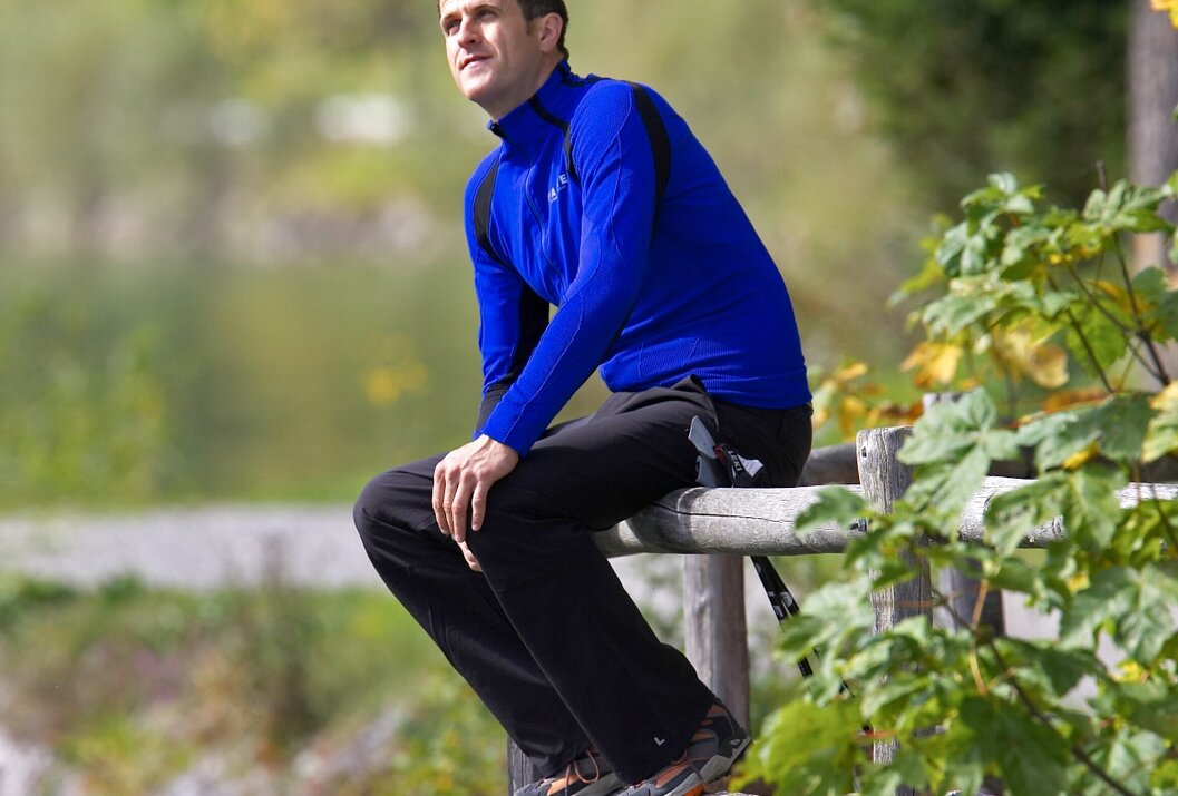 Mann in blauer Sportkleidung sitzt auf Holzzaun vor See und Bäumen