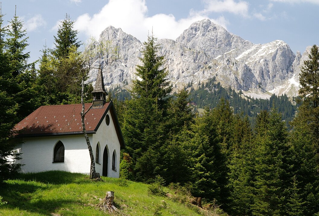 Kleine weiße Kapelle vor bewaldetem Hang mit schneebedeckten Bergen im Hintergrund