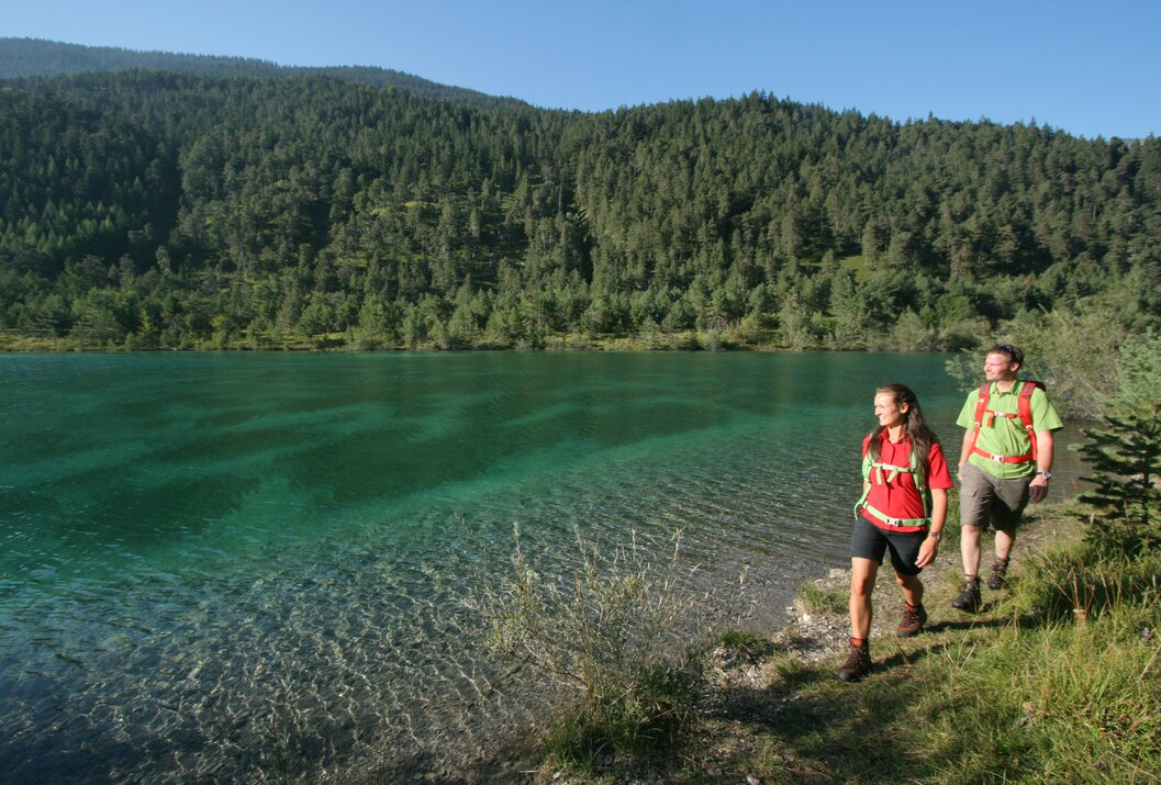 Wanderer am Ufer eines klaren Sees vor bewaldeten Bergen