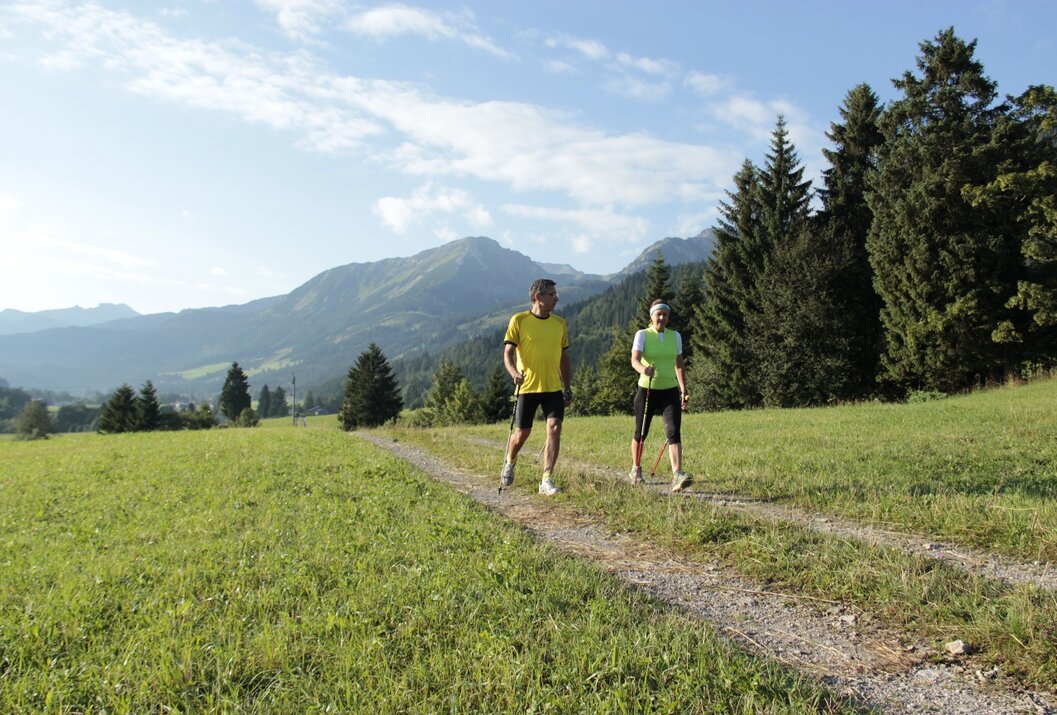 Paar wandert mit Stöcken auf einem Pfad durch eine grüne Wiese vor Bergkulisse