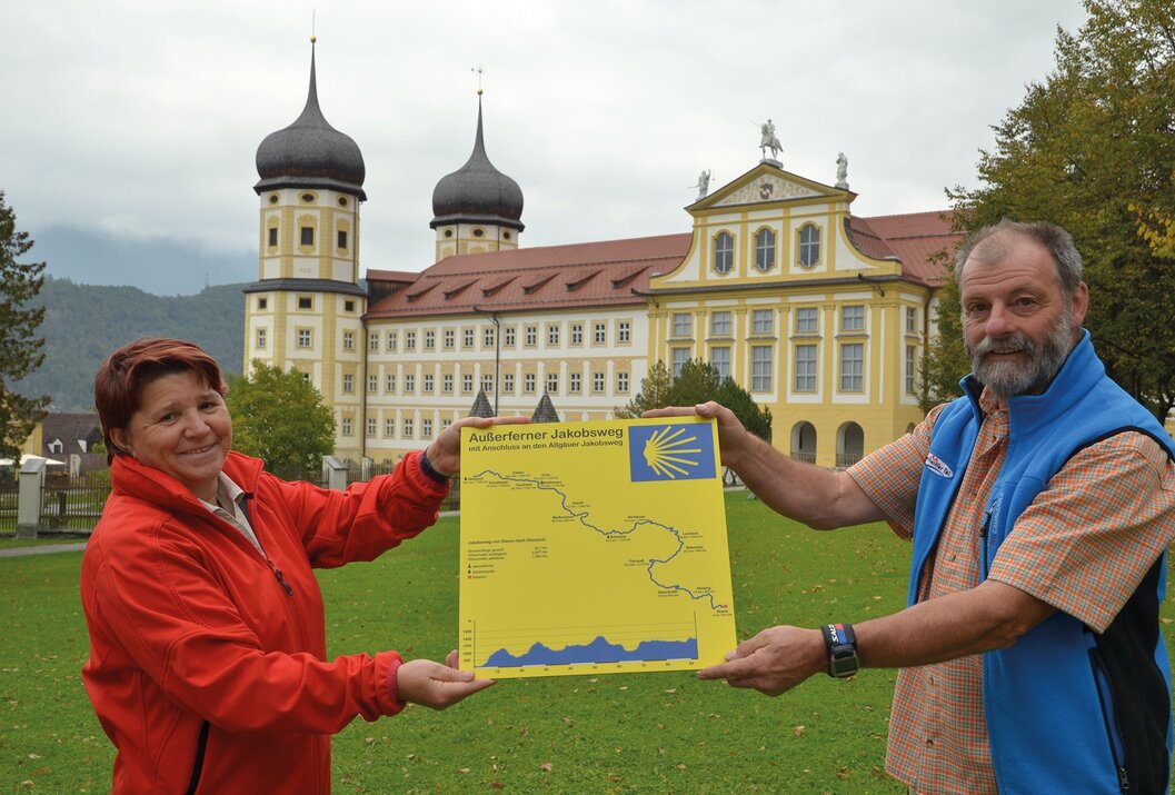 Zwei Personen halten gelbe Karte des Außerferner Jakobswegs vor Schloss mit Türmen