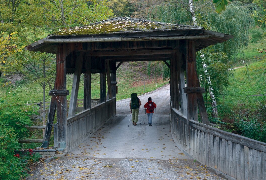 Zwei Wanderer gehen über eine überdachte Holzbrücke im Wald