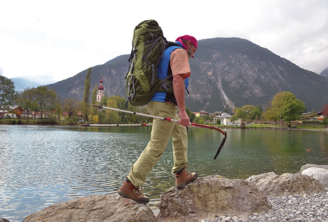Wanderer mit Rucksack und Stock überquert Steine am See vor Bergkulisse