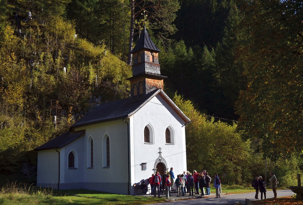 Kleine weiße Kirche mit Turm vor bewaldetem Hang, davor eine Gruppe Menschen