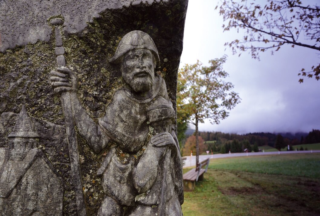 Geschnitzte Holzfigur eines Mannes mit Bart und Stock an einem Baum neben einer Bank und einer Straße