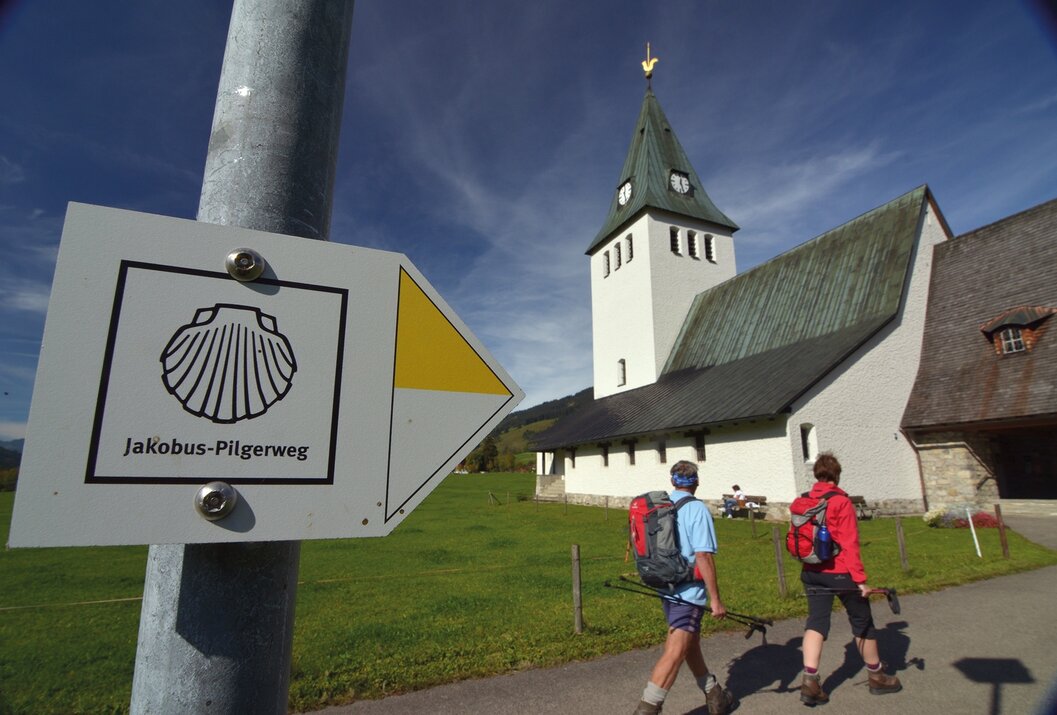 Pilgerwegweiser mit Jakobsmuschel vor Kirche und Wanderern